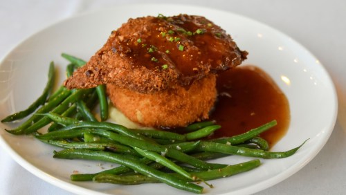A plate featuring fried chicken atop a round potato cake, drizzled with sauce, and served with green beans.