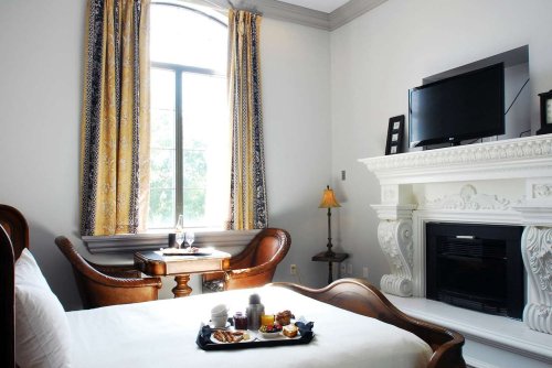 Cozy hotel room with a bed, fireplace, TV, and a table set for breakfast by a large window with elegant curtains.