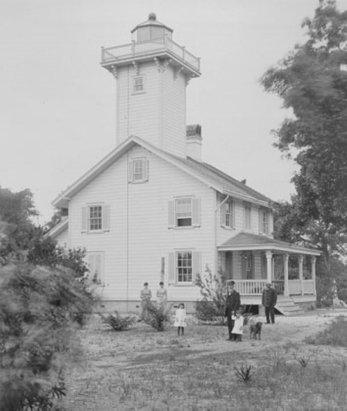 A historic white house with a lighthouse tower, featuring people and a dog in the foreground.