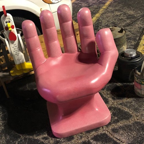 A large, pink hand-shaped chair sits on a pavement, next to a chicken statue and a can of paint.