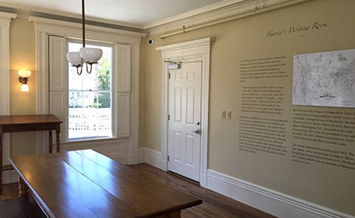 A well-lit room with a wooden table, wall text, and a window, showcasing a historical writing space.