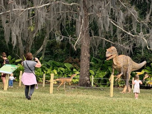 A family observes a life-sized dinosaur model in a park, surrounded by lush greenery and Spanish moss.
