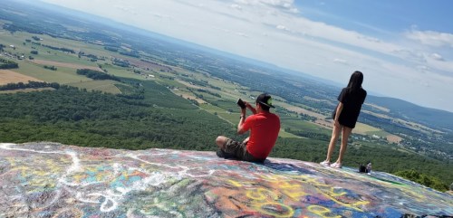 Two people on a rocky overlook, one sitting and taking photos, the other standing, with a scenic valley view below.