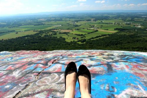 A pair of black shoes on a colorful, graffiti-covered rock ledge overlooking a lush green valley and distant hills.