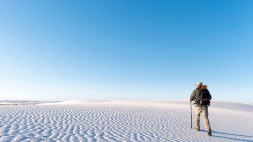 A hiker with a backpack stands on white sand dunes under a clear blue sky, gazing into the distance.