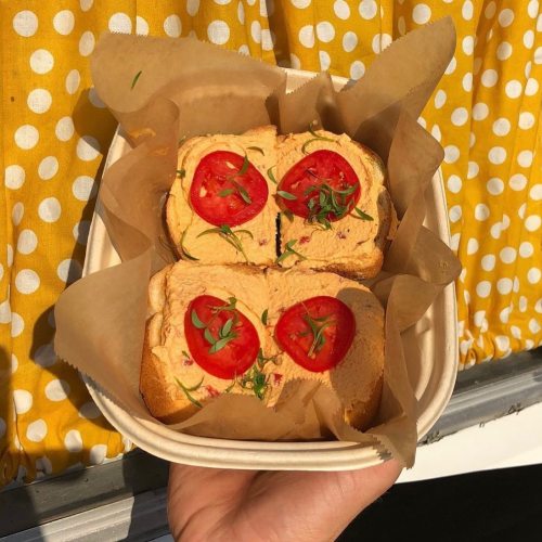 A hand holding a container with four slices of bread topped with tomato slices and herbs, set against a yellow polka dot background.