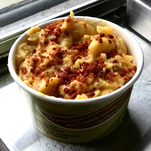 A cup of creamy macaroni and cheese topped with crispy bacon bits, sitting on a metal surface.