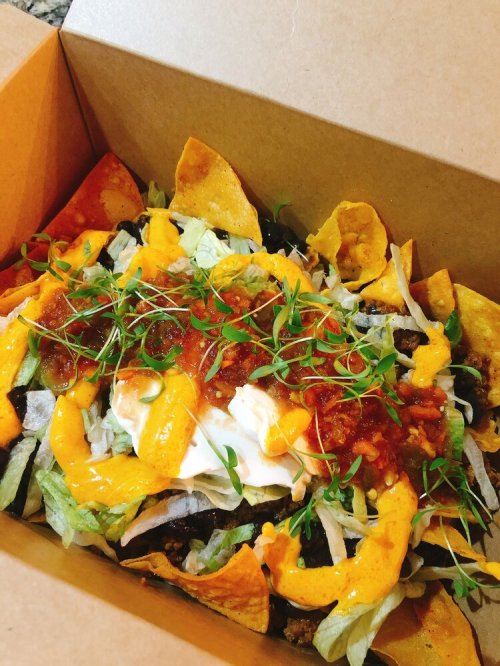 A takeout box filled with nachos topped with lettuce, cheese, sour cream, salsa, and fresh herbs.