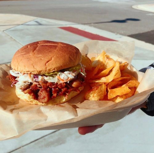 A hand holding a pulled pork sandwich topped with coleslaw, served with a side of potato chips on parchment paper.