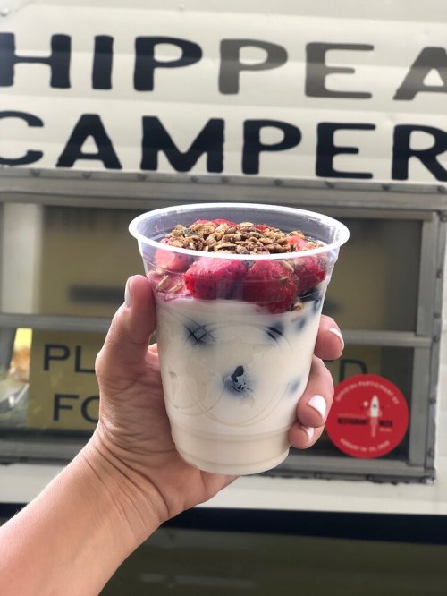 A hand holding a cup of yogurt topped with strawberries and granola, in front of a food truck.