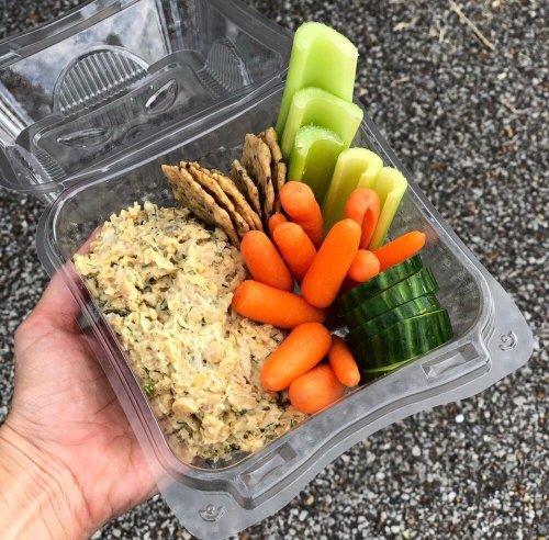 A hand holding a clear container filled with tuna salad, carrots, cucumber slices, celery, and crackers.