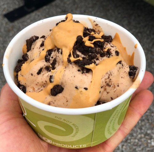 A hand holding a cup of ice cream topped with crushed cookies and a drizzle of peanut butter.