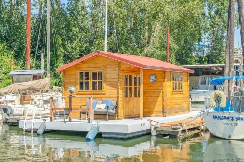A cozy wooden houseboat with a red roof, docked among sailboats in a serene marina setting.