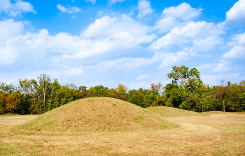 Two grassy mounds in a field under a blue sky with scattered clouds, surrounded by trees in the background.