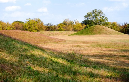 A grassy landscape with two earthen mounds and trees in the background under a partly cloudy sky.