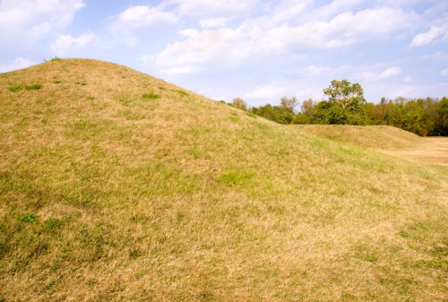 Two grassy mounds under a blue sky with scattered clouds, surrounded by trees in a natural landscape.