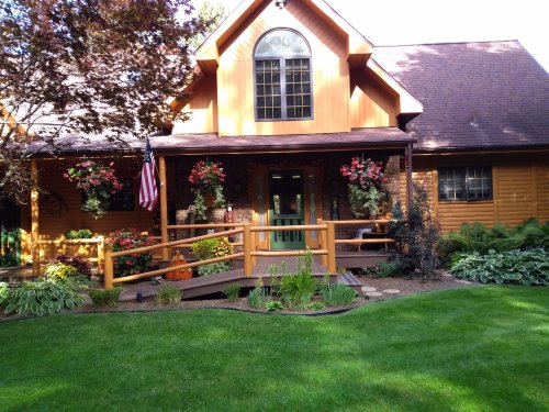 A charming wooden house with a front porch, surrounded by lush greenery and colorful flower baskets, and an American flag.