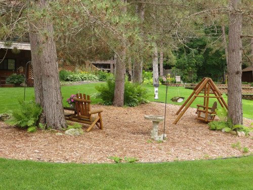 A serene garden area with wooden benches, a swing set, and lush greenery surrounded by tall trees.