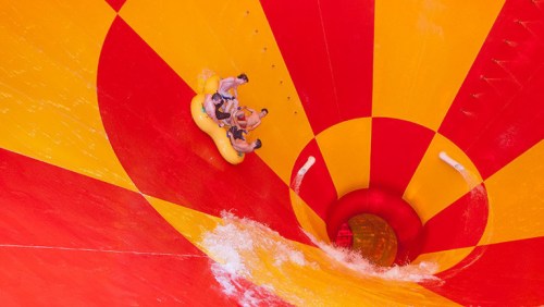 Two people on a yellow inflatable tube slide down a colorful, spiraling water slide. Water splashes around them.