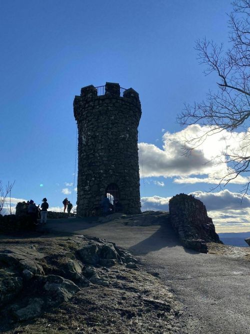 A stone tower stands against a blue sky, with people gathered nearby and clouds in the background.