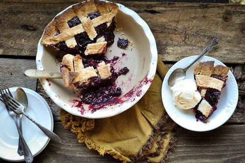 A slice of berry pie on a plate next to a scoop of ice cream, with the pie dish on a rustic wooden table.