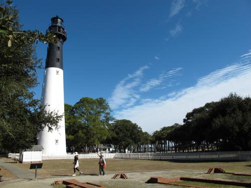 A tall lighthouse stands beside a grassy area, with two people walking and trees under a blue sky with wispy clouds.