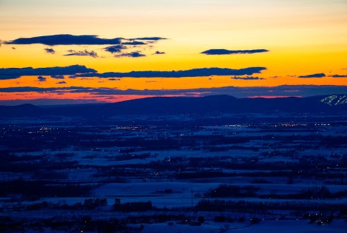A vibrant sunset over a snowy landscape, with rolling hills and distant lights twinkling in the twilight.