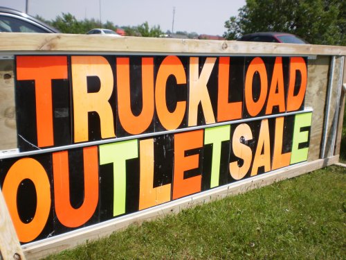Brightly colored sign reading "TRUCKLOAD OUTLET SALE" displayed outdoors on a wooden frame.