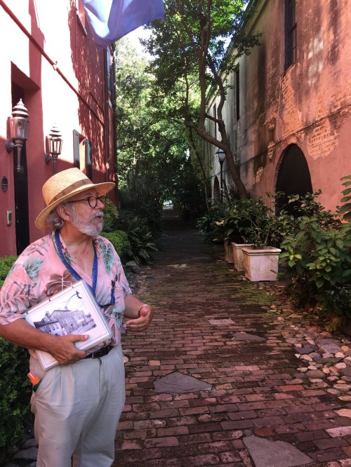 A man in a straw hat holds a photo while standing in a lush, narrow alleyway lined with greenery and historic buildings.