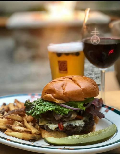 A gourmet burger with lettuce, bacon, and toppings, served with fries, a glass of beer, and a glass of red wine.