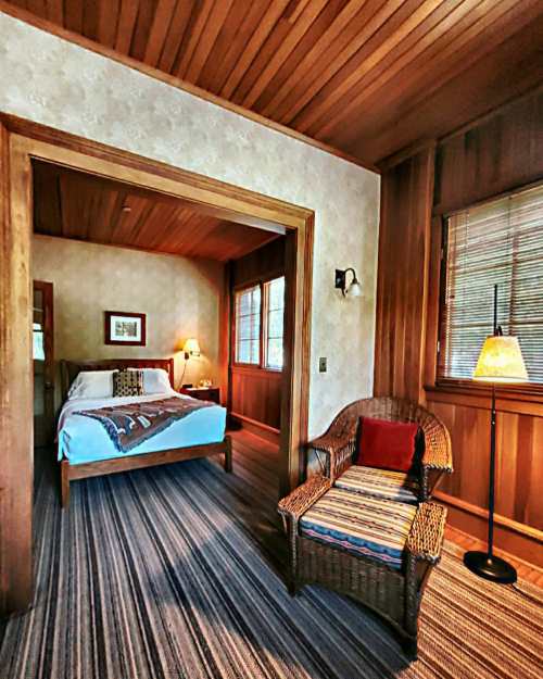 Cozy bedroom with wooden walls, a bed, a wicker chair, and a lamp, featuring warm, inviting decor.