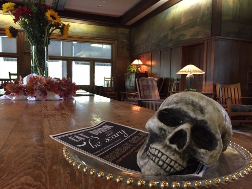 A decorative skull on a table with autumn leaves, a sign, and flowers in a cozy, wooden room.