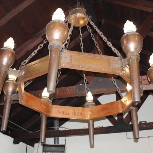 A vintage chandelier with eight candle-like lights, hanging from a wooden ceiling with dark beams.