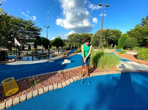 A woman in a green shirt plays mini-golf on a blue course surrounded by greenery and a pond under a sunny sky.