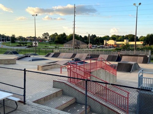 A skate park featuring ramps, rails, and a concrete area, surrounded by a fence and grassy fields in the background.