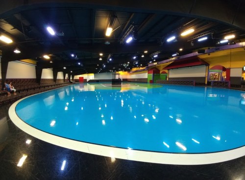 A spacious indoor skating rink with a bright blue floor and colorful walls, featuring seating along the edges.