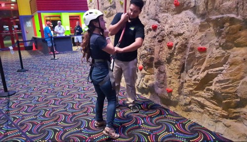 A person in a helmet prepares to climb a rock wall with an instructor's assistance in a colorful indoor climbing facility.