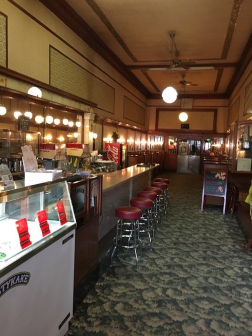 Interior of a vintage diner with a long counter, red stools, and retro decor, featuring a soda fountain and patterned carpet.