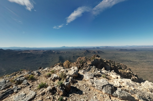 Panoramic view from a rocky mountain peak, showcasing vast desert landscapes and distant mountains under a clear blue sky.