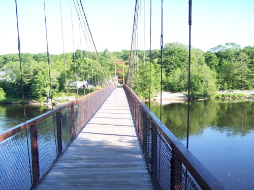 A wooden suspension bridge spans a calm river, surrounded by lush green trees under a clear blue sky.