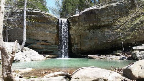 A serene waterfall cascades into a clear pool, surrounded by rocky cliffs and lush greenery.