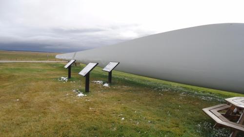 A large wind turbine blade rests on the ground, with informational signs nearby in a grassy field under a cloudy sky.