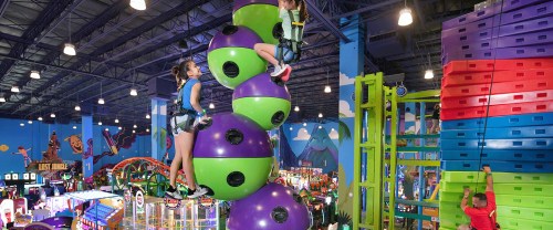 Two children climb a colorful, stacked climbing structure in a vibrant indoor amusement park filled with games and attractions.