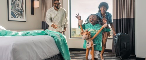 A joyful girl in a green shirt runs with a stuffed giraffe, while her parents smile in a hotel room.