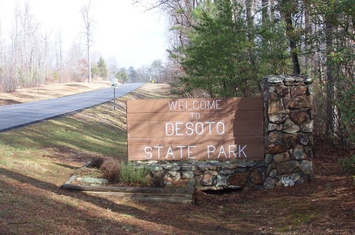 A wooden sign reading "Welcome to Desoto State Park" beside a road, surrounded by trees and greenery.