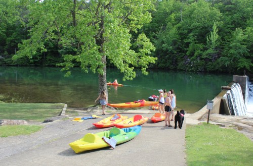 A group of people by a calm lake with kayaks, a waterfall, and lush green trees in the background.