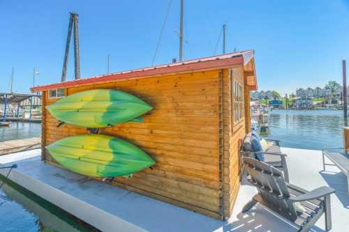 A wooden shed at a marina with two green kayaks mounted on the side, surrounded by water and boats.