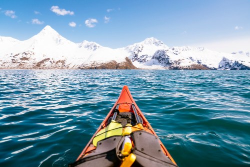 A kayak glides over calm waters, with snow-capped mountains rising in the background under a clear blue sky.