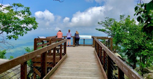 Three people stand on a wooden observation deck, overlooking a scenic landscape with blue skies and distant mountains.