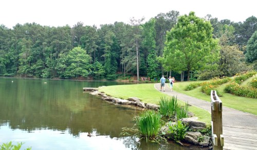 A serene lake surrounded by greenery, with people walking along a path beside the water.